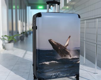 May include: A black suitcase with a printed image of a humpback whale breaching in the ocean. The image is signed "Tim Stevens Photography".