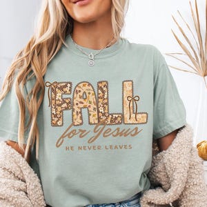 May include: A light green t-shirt with the words "FALL for Jesus" in a floral design. The text "HE NEVER LEAVES" is below in a brown script. The shirt has short sleeves and a relaxed fit.