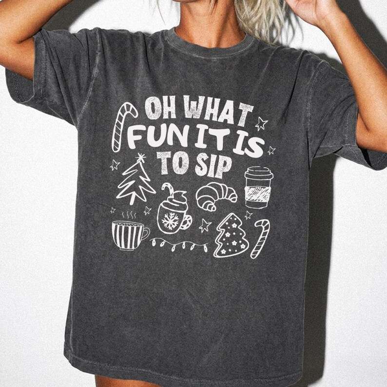 May include: A charcoal grey short-sleeved t-shirt featuring white screen-printed graphics of holiday icons including a Christmas tree, a mug, a croissant, and a coffee cup, with the text "OH WHAT FUN IT IS TO SIP" centred on the chest.