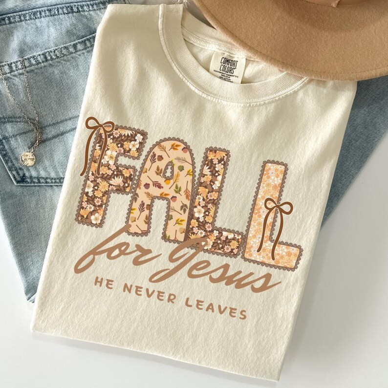 May include: Cream-colored t-shirt with the words "FALL for Jesus" in a floral design. The shirt also has the text "HE NEVER LEAVES" in a brown script. A tan hat and blue jeans are in the background.