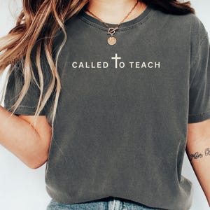 May include: A dark grey t-shirt with the text "CALLED TO TEACH" and a small cross graphic printed on the front.