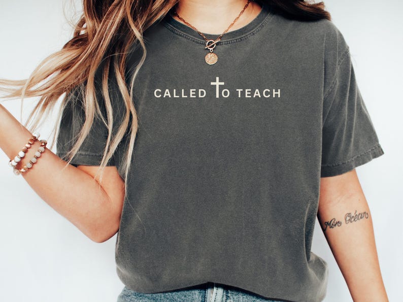 May include: A dark grey t-shirt with the text "CALLED TO TEACH" and a small cross graphic printed on the front.