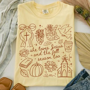 May include: A light yellow t-shirt with a crew neck and short sleeves. The shirt features a brown graphic design with a cross, flowers, a pumpkin, and the text "she loves Jesus and the fall season too."