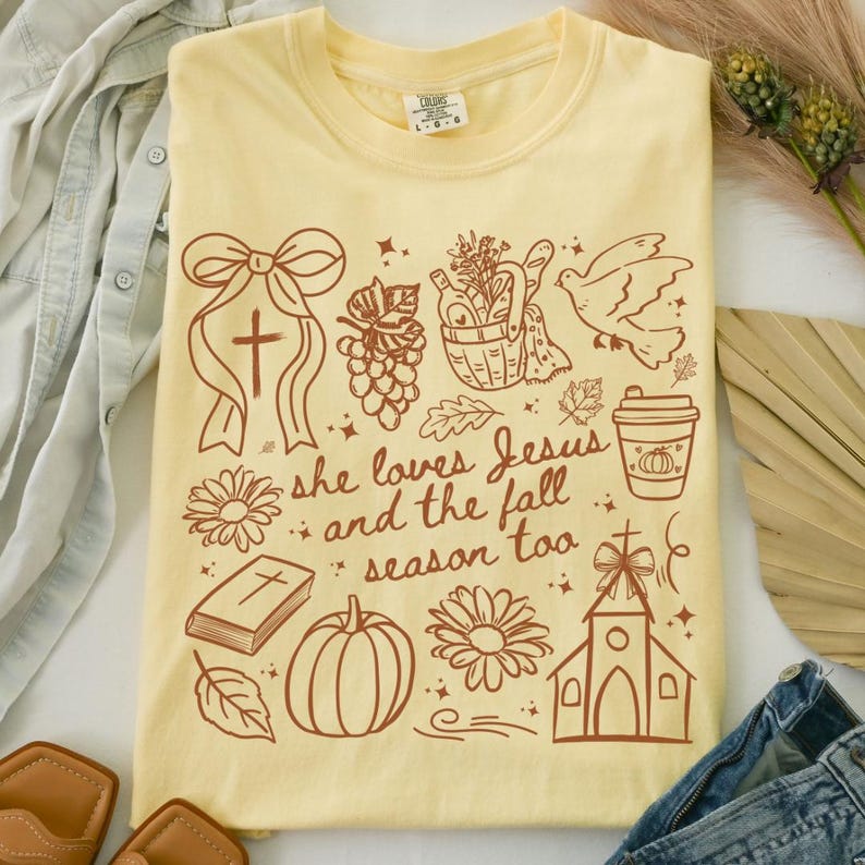 May include: A light yellow t-shirt with a crew neck and short sleeves. The shirt features a brown graphic design with a cross, flowers, a pumpkin, and the text "she loves Jesus and the fall season too."