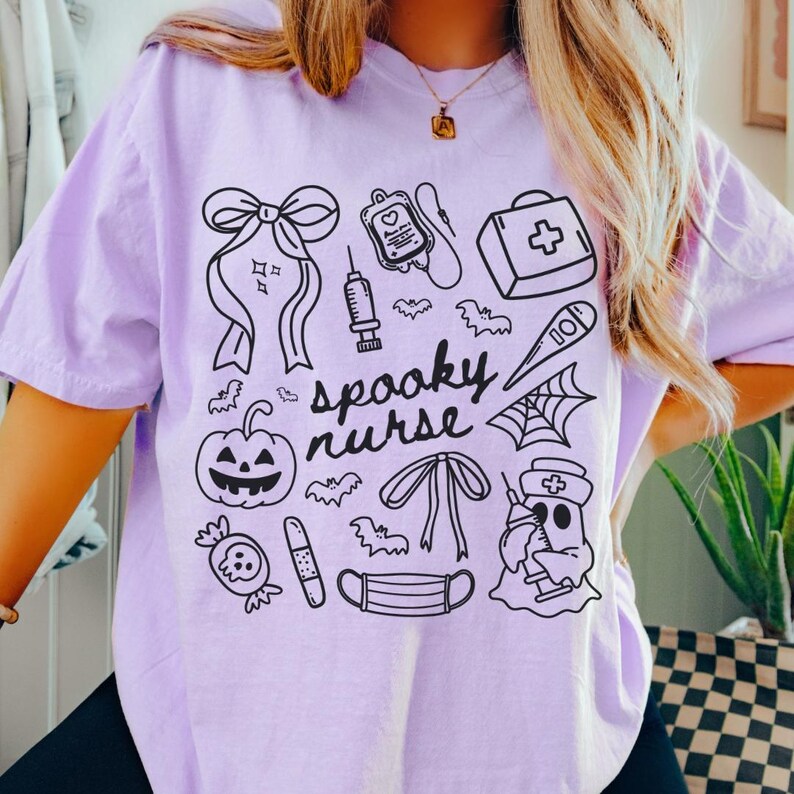 以下が含まれることがあります： 黒と白のグラフィックデザインが施された、薄紫色のTシャツ。デザインには、カボチャ、コウモリ、注射器、そして「spooky nurse」という言葉など、ハロウィンと医療をテーマにしたイラストが含まれています。
