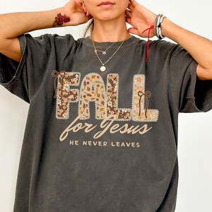 May include: Dark gray t-shirt with the words "FALL for Jesus" in a floral design. The text "HE NEVER LEAVES" is below the main text. The letters are outlined with a decorative border.