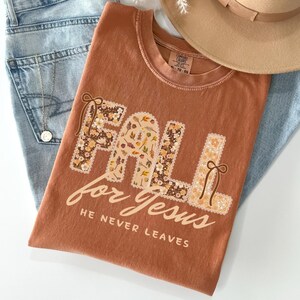 May include: A rust-colored t-shirt with the words "FALL for Jesus" in a floral design. The shirt is laid on top of blue jeans and a tan hat. The text "HE NEVER LEAVES" is printed below the main text.