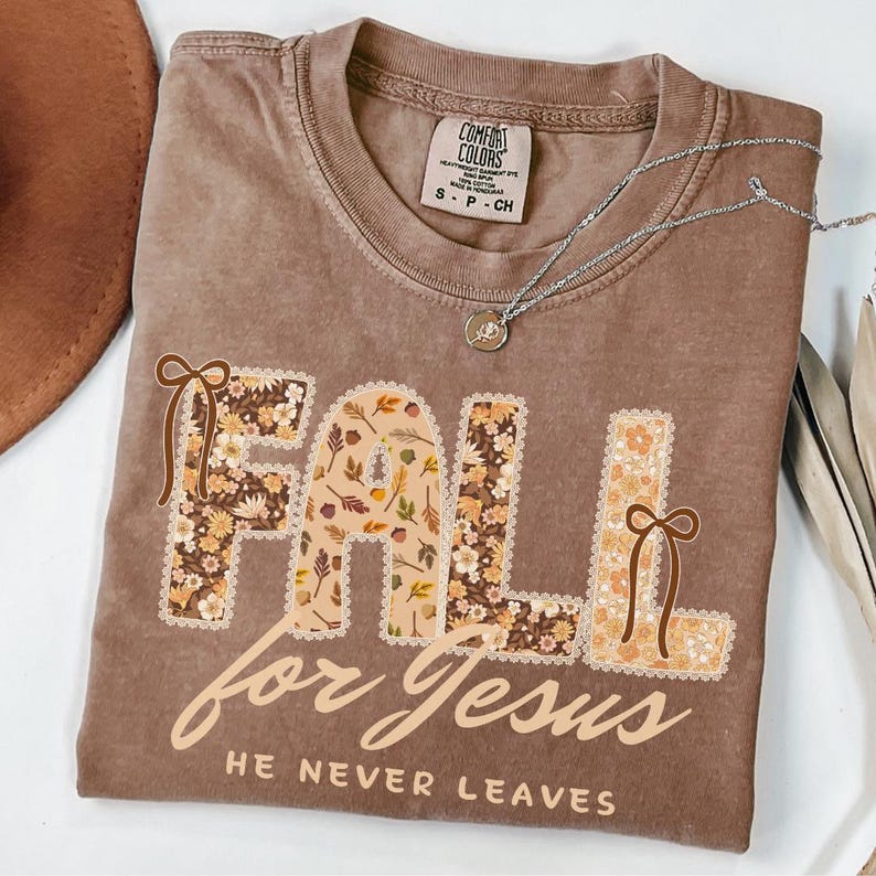 May include: A brown Comfort Colors t-shirt with the words "FALL for Jesus" in a floral design. The text "HE NEVER LEAVES" is below. A silver necklace with a pendant is on the shirt.