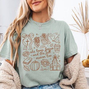 May include: A light green t-shirt with a brown graphic design featuring a cross, flowers, a pumpkin, a church, and the text "she loves Jesus and the fall season too."