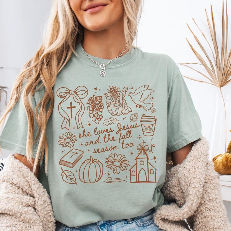 May include: A light green t-shirt with a brown graphic design featuring a cross, flowers, a pumpkin, a church, and the text "she loves Jesus and the fall season too."
