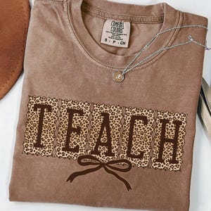 May include: A brown t-shirt with the word "TEACH" in a leopard print design. The shirt has a crew neck and short sleeves. A silver necklace with a small pendant is on the shirt.