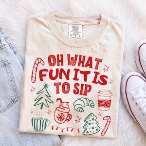 May include: A beige short-sleeve t-shirt featuring red and green holiday-themed graphics and the text "OH WHAT FUN IT IS TO SIP". The design includes a candy cane, a Christmas tree, a coffee cup, a croissant, and a mug with a snowflake pattern.