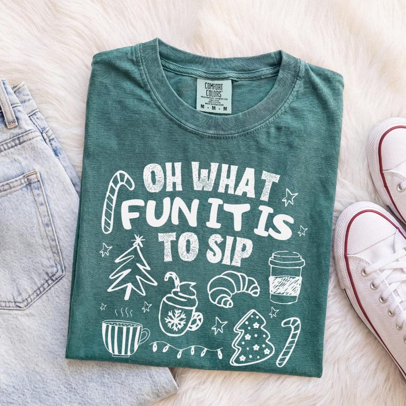 May include: A dark green t-shirt featuring white graphic illustrations of a candy cane, a Christmas tree, a mug with a snowflake, a croissant, a coffee cup, and a decorated tree, with the text "OH WHAT FUN IT IS TO SIP" printed in white.