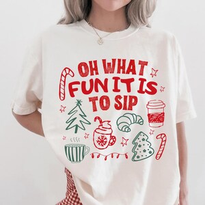 May include: A cream-coloured short-sleeve t-shirt featuring red and green holiday-themed graphics and the text "OH WHAT FUN IT IS TO SIP". The design includes illustrations of a candy cane, a Christmas tree, a coffee cup, a croissant, and stars.