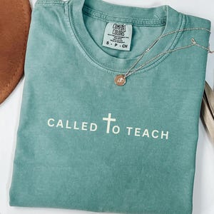 May include: A teal blue t-shirt with the text "CALLED TO TEACH" and a white cross in the centre. The shirt is made of a soft, comfortable fabric and has a distressed look.