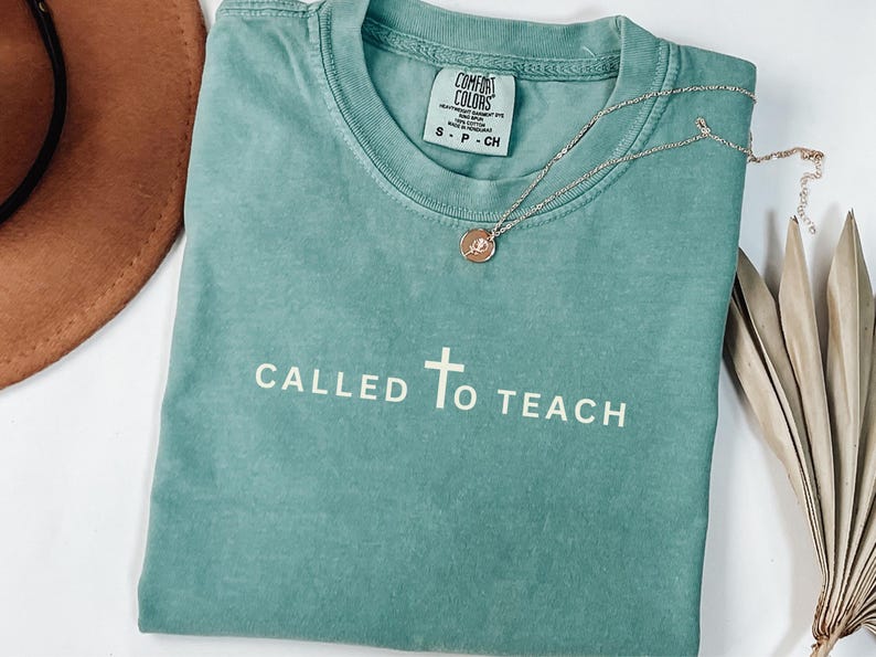 May include: A teal blue t-shirt with the text "CALLED TO TEACH" and a white cross in the centre. The shirt is made of a soft, comfortable fabric and has a distressed look.