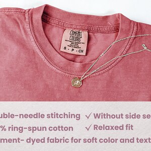 May include: A close-up of a dusty rose coloured t-shirt with a tag that reads "Comfort Colors" and "S-P-CH". The t-shirt is made of 100% ring-spun cotton and features double-needle stitching, a relaxed fit, and garment-dyed fabric for a soft colour and texture.