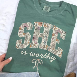 May include: A green t-shirt with a floral print design that spells out "SHE is worthy" with a bow underneath.