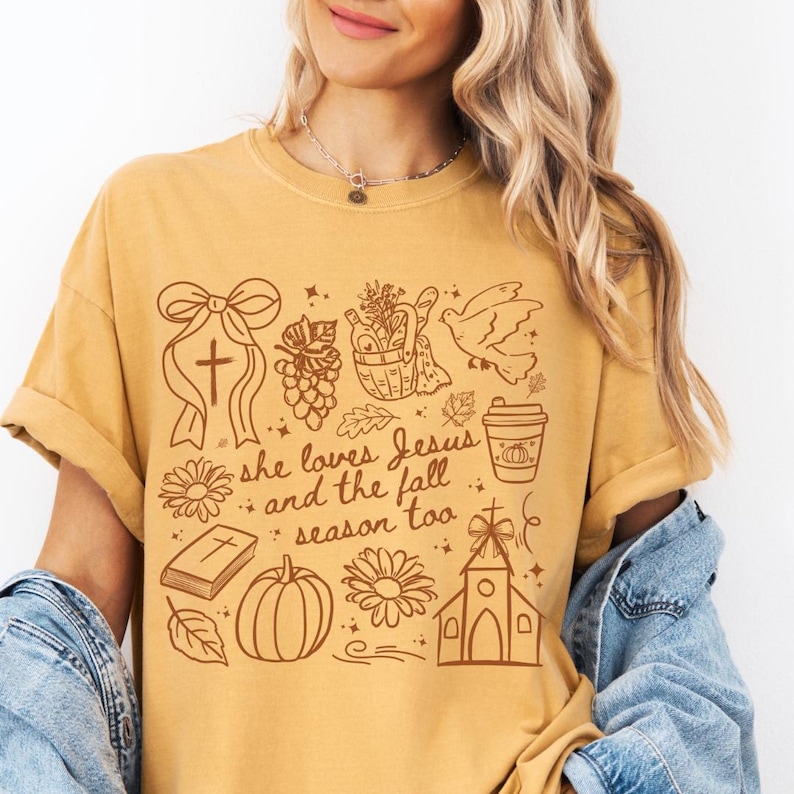 May include: Mustard yellow t-shirt with a brown graphic design. The design includes a cross, grapes, a dove, a coffee cup, a pumpkin, a church, and the text "she loves Jesus and the fall season too."