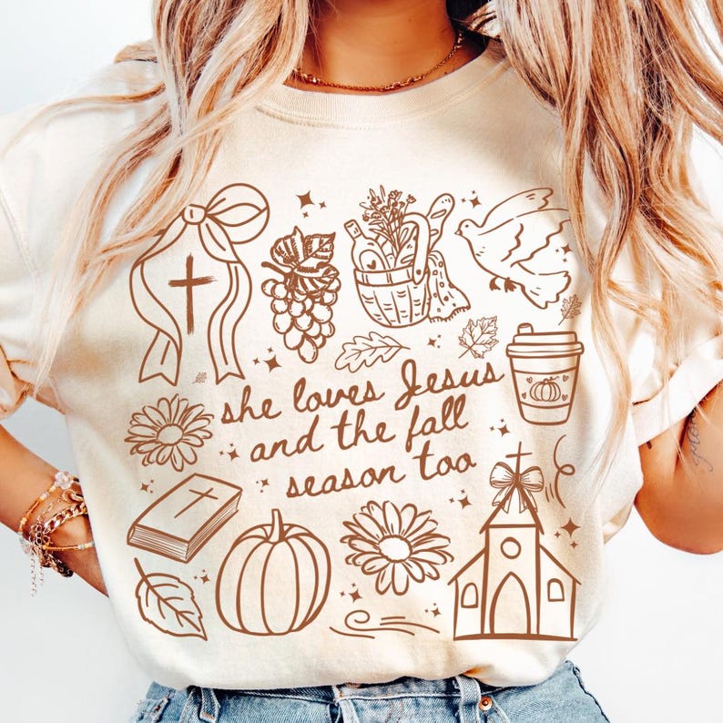 May include: Cream-colored t-shirt with a brown graphic design. The design includes a cross, a dove, a pumpkin, a church, and the text "she loves Jesus and the fall season too."