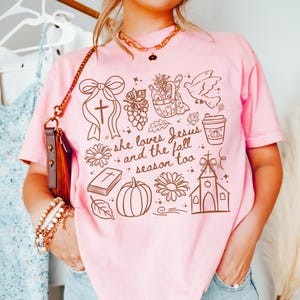 May include: Pink t-shirt with a brown graphic design featuring a cross, flowers, a pumpkin, and the text "she loves Jesus and the fall season too." The shirt is a light pink color.