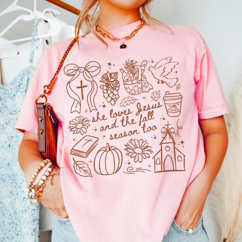 May include: Pink t-shirt with a brown graphic design featuring a cross, flowers, a pumpkin, and the text "she loves Jesus and the fall season too." The shirt is a light pink color.