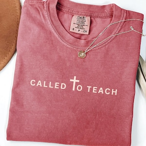 May include: A red t-shirt with the words "CALLED TO TEACH" and a cross printed on the front. The shirt is made by Comfort Colors.