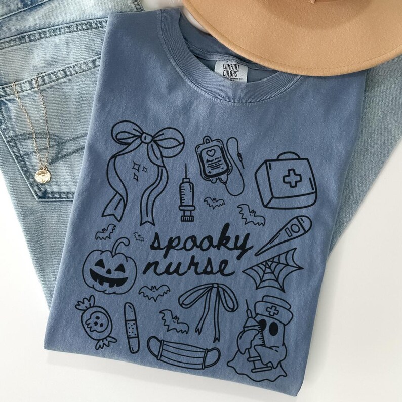 以下が含まれることがあります： ハロウィーンをテーマにした医療シンボルと「spooky nurse」の文字が描かれた黒いグラフィックデザインの青いComfort Colors Tシャツ。デザインには、カボチャ、コウモリ、注射器、救急箱が含まれています。