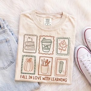 May include: A beige t-shirt featuring a graphic with six rectangular frames containing line art of books, a coffee cup, a leaf, a pumpkin, a pencil holder, and a pencil. The text "FALL IN LOVE WITH LEARNING" is printed in brown below the frames.