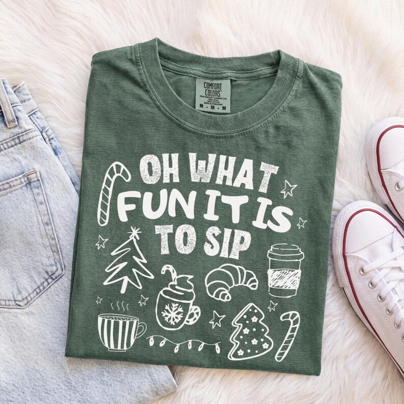 May include: A dark green t-shirt featuring white graphic illustrations of a candy cane, a Christmas tree, a mug, a croissant, and a coffee cup. The text reads "OH WHAT FUN IT IS TO SIP". The shirt has a crew neckline and a size M label at the collar.
