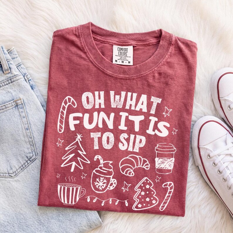 May include: A maroon short-sleeved t-shirt featuring white graphic text that reads "OH WHAT FUN IT IS TO SIP" surrounded by illustrations of a candy cane, a Christmas tree, a mug, a croissant, a coffee cup, and a decorated cookie.