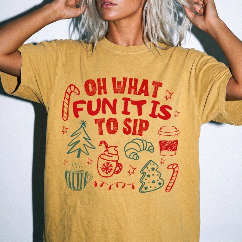 May include: A mustard yellow short-sleeve t-shirt featuring red text that reads OH WHAT FUN IT IS TO SIP. The graphic includes red and green illustrations of a candy cane, a Christmas tree, a mug, a croissant, a coffee cup, and stars.