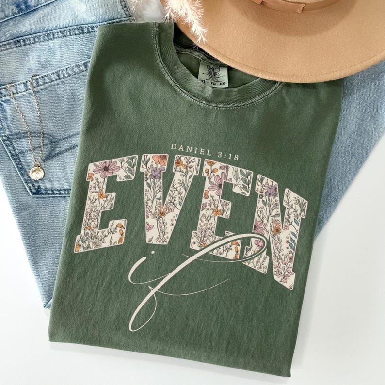 Puede incluir: Camiseta verde oliva con la palabra "EVEN" en un dise&ntilde;o floral, con "Daniel 3:18" arriba. La camiseta est&aacute; colocada sobre unos vaqueros azules, un collar dorado y un sombrero beige.