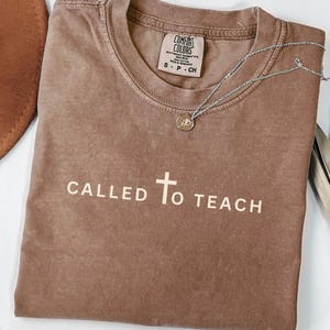 May include: A brown t-shirt with the text "CALLED TO TEACH" and a cross in white. The shirt has a distressed look and is made of a soft, comfortable fabric.