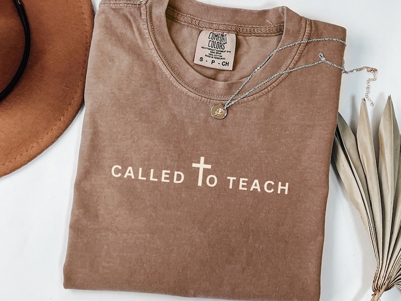 May include: A brown t-shirt with the text "CALLED TO TEACH" and a cross in white. The shirt has a distressed look and is made of a soft, comfortable fabric.
