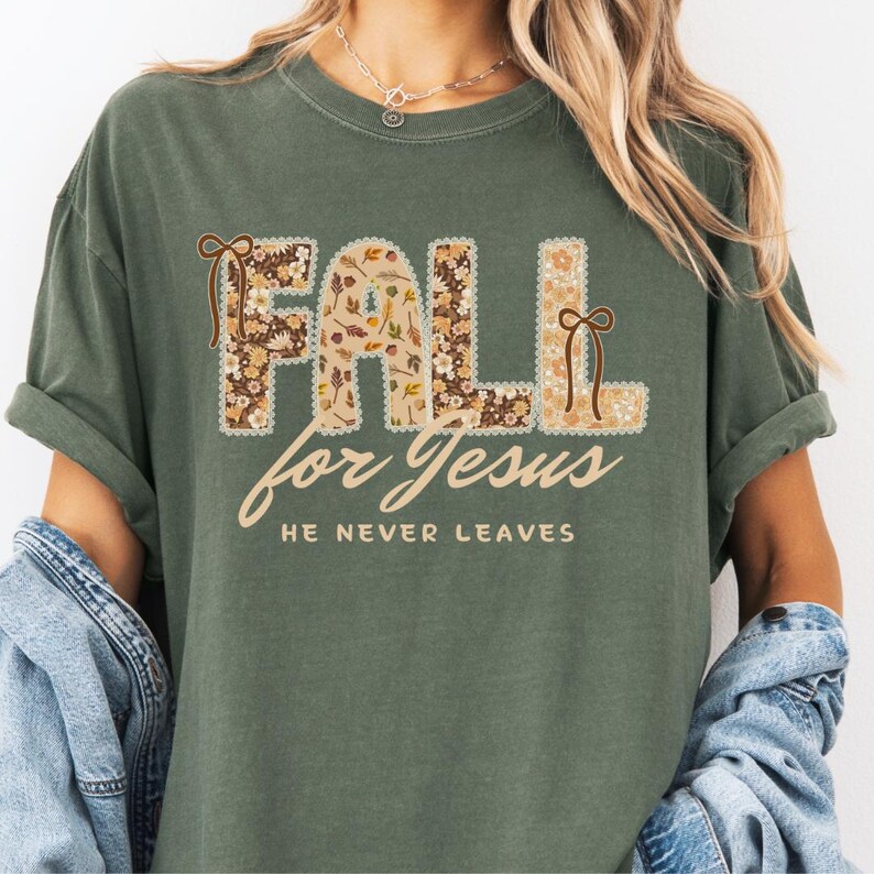 May include: Olive green t-shirt with the words "FALL for Jesus" in a floral design. The text "HE NEVER LEAVES" is below in a script font. The shirt has rolled-up sleeves.