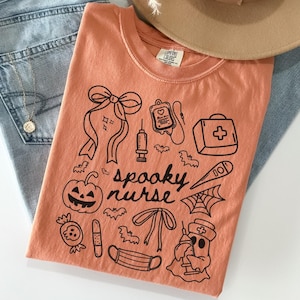 以下が含まれることがあります： オレンジ色のTシャツ。黒いグラフィックデザインで、カボチャ、コウモリ、医療用品、そして「spooky nurse」の文字が描かれています。Tシャツはブルージーンズとベージュの帽子の上に折りたたまれています。