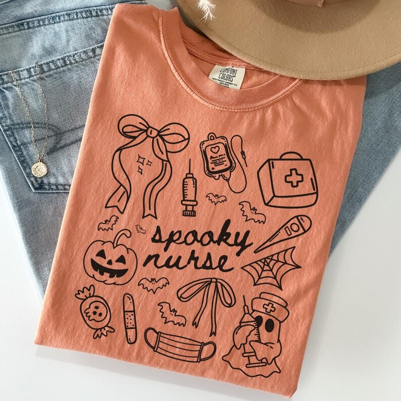 以下が含まれることがあります： オレンジ色のTシャツ。黒いグラフィックデザインで、カボチャ、コウモリ、医療用品、そして「spooky nurse」の文字が描かれています。Tシャツはブルージーンズとベージュの帽子の上に折りたたまれています。