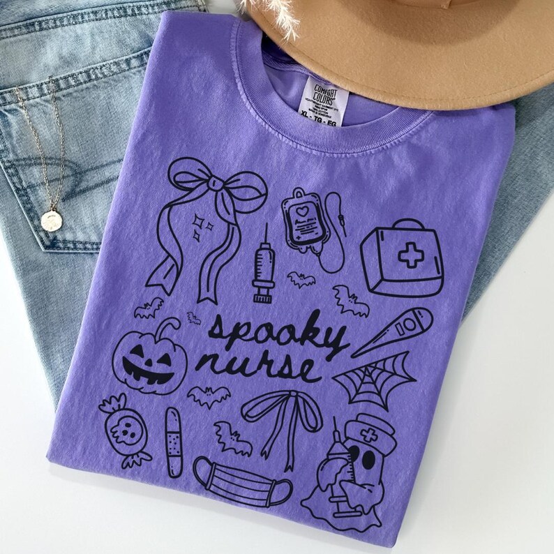 以下が含まれることがあります： ハロウィーンをテーマにした医療イラストと「spooky nurse」の文字が描かれた黒いグラフィックデザインの紫色のComfort Colors Tシャツ。デザインにはカボチャ、コウモリ、医療器具が含まれています。