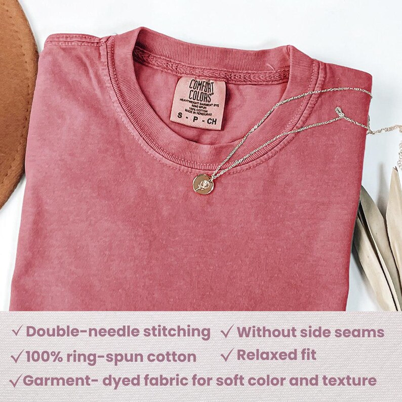 May include: A dusty rose Comfort Colors t-shirt with a crew neck, folded on a surface. A silver necklace with a circular pendant rests on the shirt. The shirt tag reads "COMFORT COLORS". Features include double-needle stitching and ring-spun cotton.