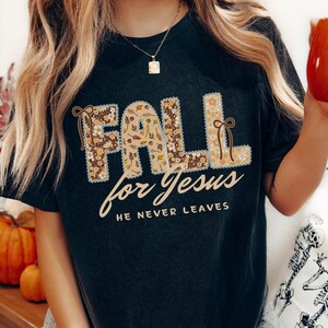 May include: Dark gray t-shirt with the words "FALL for Jesus" in a floral design. The text "HE NEVER LEAVES" is below the main text. The shirt has a round neckline and short sleeves.
