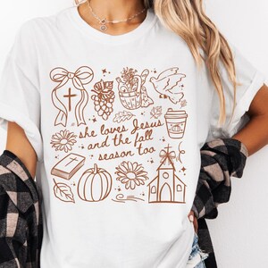 May include: White t-shirt with a brown graphic design featuring a cross, grapes, a dove, a pumpkin spice latte, a pumpkin, a church, and the text "she loves Jesus and the fall season too."