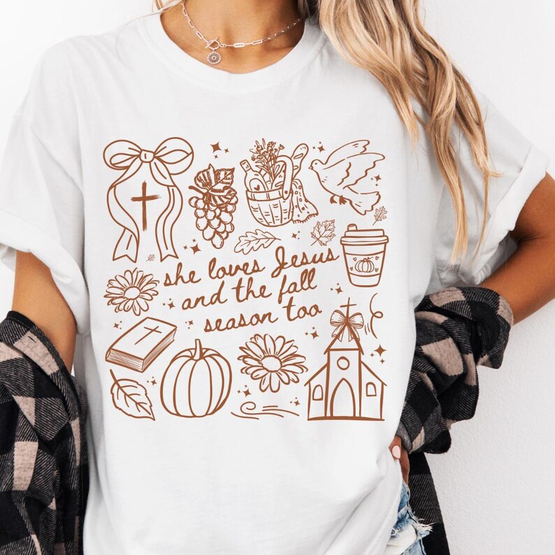May include: White t-shirt with a brown graphic design featuring a cross, grapes, a dove, a pumpkin spice latte, a pumpkin, a church, and the text "she loves Jesus and the fall season too."