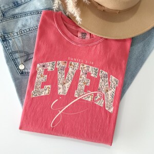 Puede incluir: Una camiseta de color coral con el texto "EVEN" en un dise&ntilde;o floral y el texto "Daniel 3:18". La camiseta est&aacute; doblada sobre unos vaqueros azules, con un sombrero beige y un collar dorado visibles.