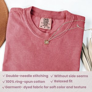 May include: A dusty rose Comfort Colors t-shirt with a crew neck, folded on a surface. A silver necklace with a circular pendant rests on the shirt. The shirt tag reads "COMFORT COLORS". Features include double-needle stitching and ring-spun cotton.