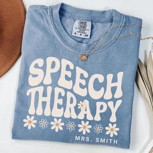 May include: A blue Comfort Colors t-shirt with the words "SPEECH THERAPY" in a retro font, decorated with daisy graphics. The shirt also includes the name "MRS. SMITH" at the bottom. A gold necklace with a pendant rests on the shirt.