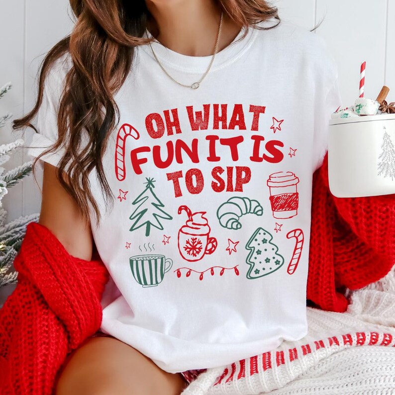 May include: A white t-shirt featuring red and green holiday-themed graphics and the text "OH WHAT FUN IT IS TO SIP". The design includes illustrations of a candy cane, a Christmas tree, a mug with whipped cream, a croissant, and a coffee cup.