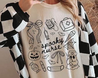 May include: Cream-colored long-sleeved shirt with a black and white checkered pattern on the sleeves. The shirt features a Halloween-themed graphic with the words "spooky nurse" and various medical and spooky illustrations.