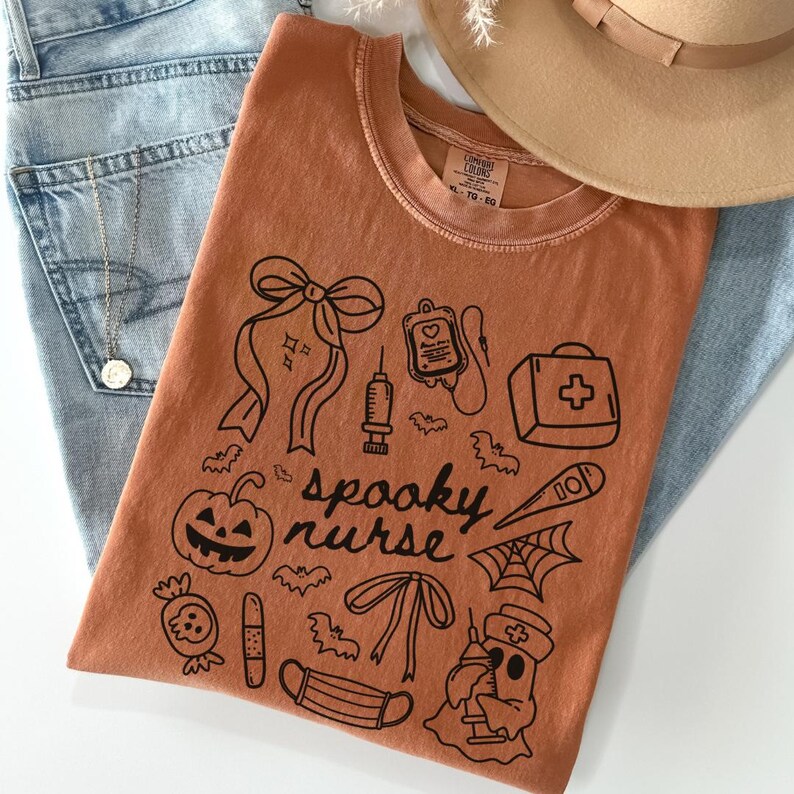 以下が含まれることがあります： ハロウィーンをテーマにした医療イラストと「spooky nurse」の文字が描かれた黒いグラフィックデザインのオレンジ色のComfort Colors Tシャツ。デザインにはカボチャ、コウモリ、幽霊の看護師が含まれています。