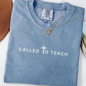 May include: A light blue t-shirt with the text "CALLED TO TEACH" and a white cross in the centre. The shirt is made of a soft, comfortable fabric and has a distressed look.